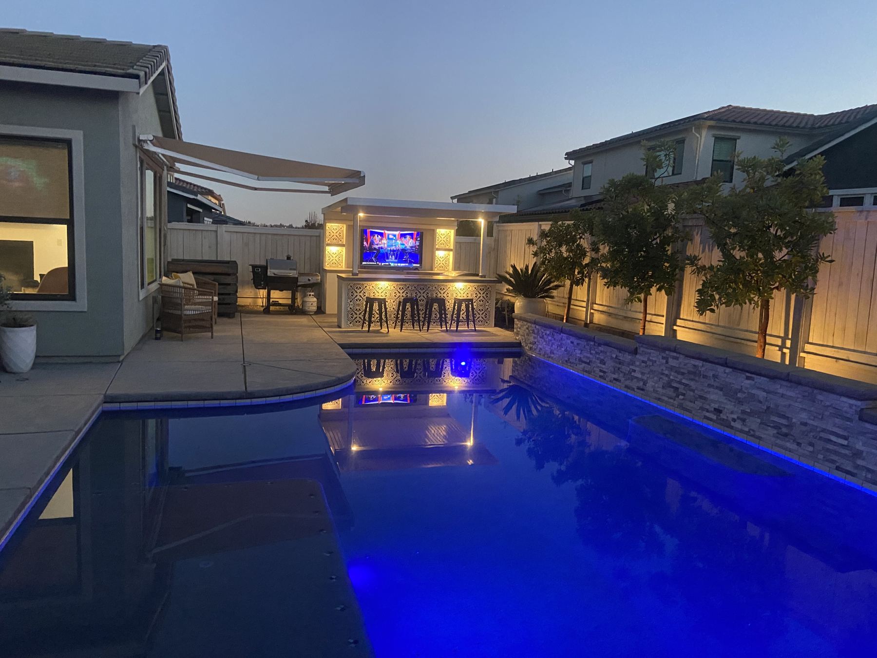Backyard at night with a lit TV island across a glowing blue pool
