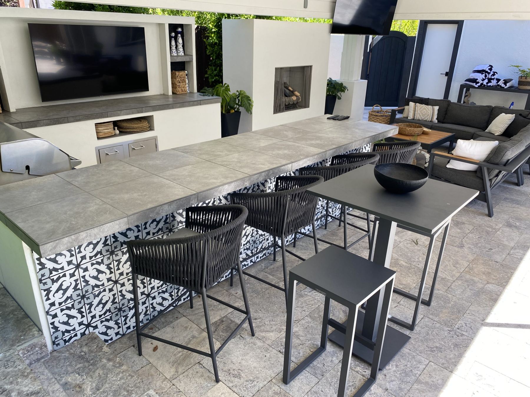 Long tiled bar with black stools, TV wall and fireplace under a covered patio